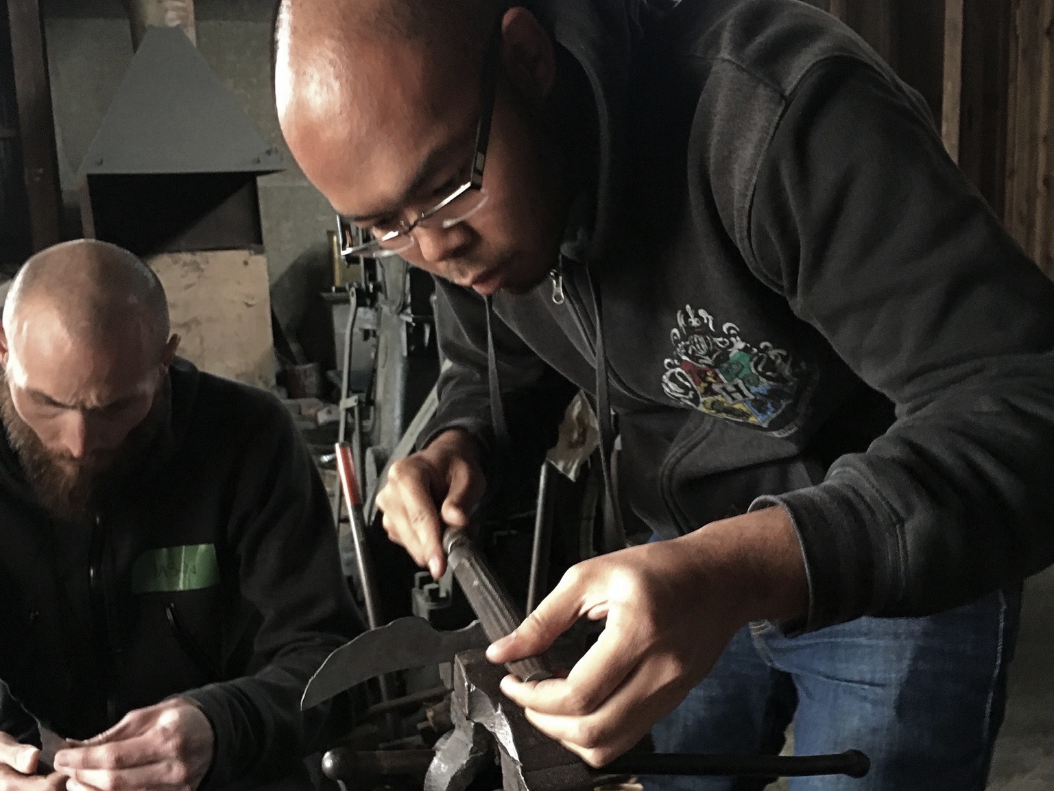 Two men are filing the sharp edges of their knife handles with a vise and a rasp.